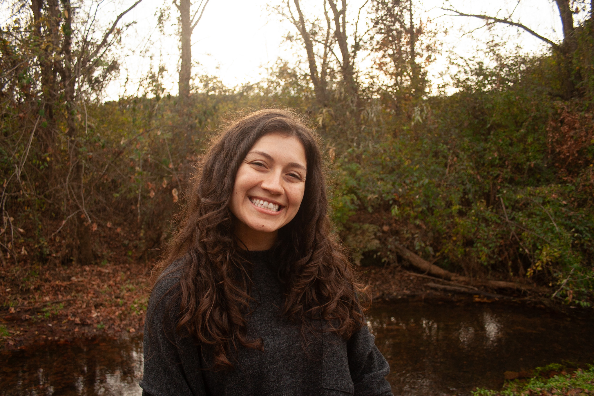 Curly brown haired woman smiling, Grey sweater,Fall tree line background Photo credit: Ryan Kirst